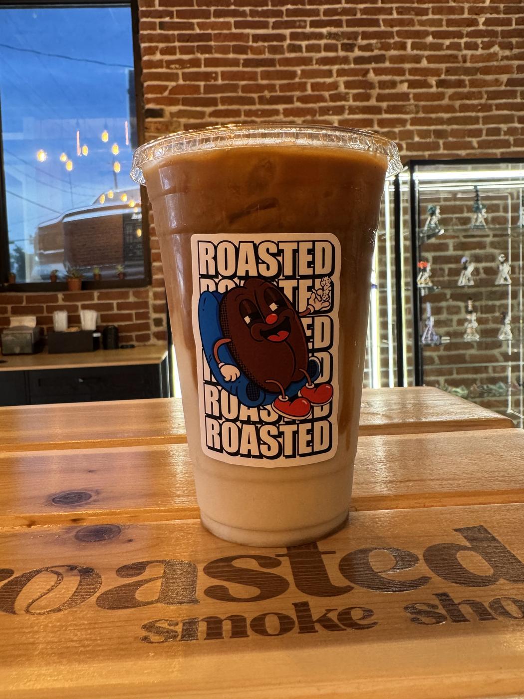 An iced coffee in a roasted. cup on the counter, the shop's name burned into the wood beside it.